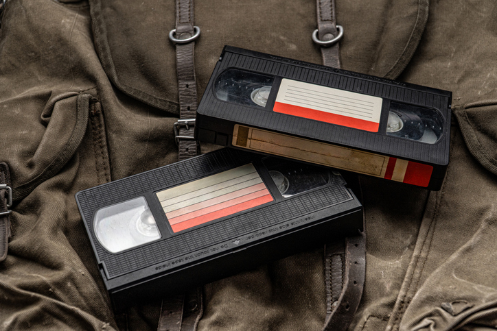 Two old black VHS cassettes with a blank label, resting on a brown canvas bag with leather straps.