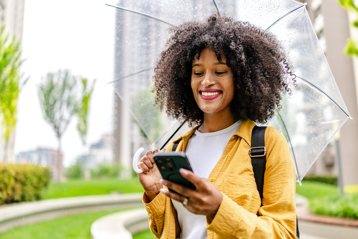 Young black woman smiles while using her phone under a clear umbrella in an urban park setting on a rainy day